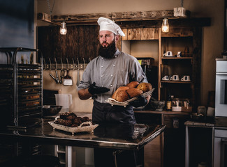 Handsome bearded chef in uniform showing tray of fresh bread in the kitchen of bakery.