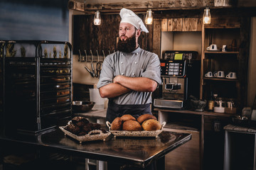 Handsome bearded chef in uniform showing tray of fresh bread in the kitchen of bakery.