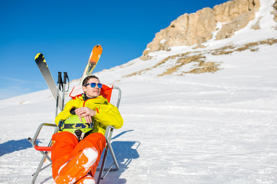 Image of sports man sitting on chair next to skis and sticks on background of snowy mountains