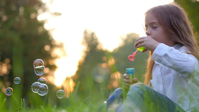 Cute little girl in a white shirt sitting on the grass in the evening park and blowing bubbles, childhood
