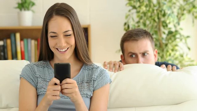 Cheater woman dating on line with a smart phone and boyfriend is spying sitting on a sofa at home