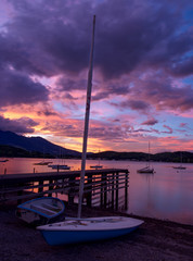 夕焼けの湖畔にて、ヨットのマストが空に聳え立つ、ボートが陸揚げして、桟橋が伸びている、湖面には数台のヨットが係留、湖面と空の厚い雲が夕焼け色に染まる。