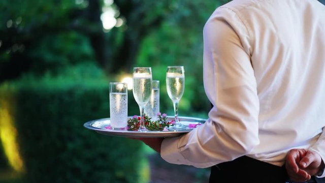 Waiter and champagne and water glasses on a tray in restaurant or event party meets clients and guests at the entrance. Catering staff. 4K UHD.