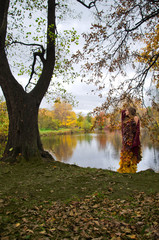 forest fairy in a dress of autumn leaves cherry-colored woolen shawl stands on the shore of a forest lake near a huge old tree