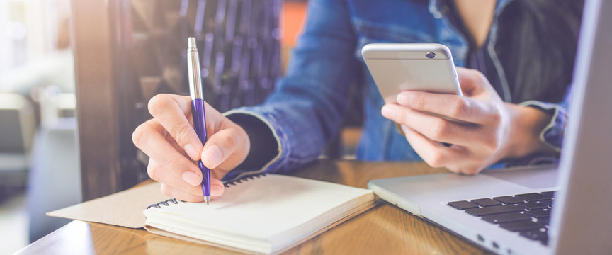 A woman's hand is writing on a notepad with a pen and a phone with a laptop.Web banner. - Powered by Adobe