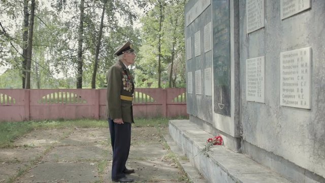 An elderly gray-haired veteran of the great Patriotic war and world war II in uniform with many badges and orders stands near the mass grave. The grandfather remembers horrors of military operations