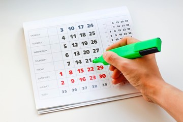 Hand crosses off the green marker days in the calendar. White calendar. Weekends are highlighted in...