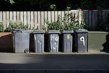 Black wheelie bins in a row on street with house numbers printed on front waiting for bin men to...