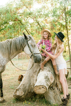 Beautiful young girl with blond hair in a suede jacket with fringe with little sister in a straw hat and checkered vintage dress with a horse in the countryside on a sunny autumn day
