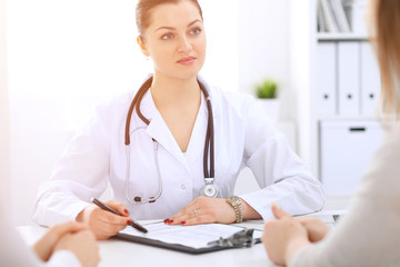 Brunette female doctor talking to patient at hospital office. Physician says about medical exams...