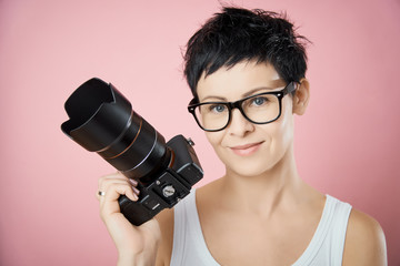 beautiful woman in glasses and with short hair with a camera in hand poses on a pink background