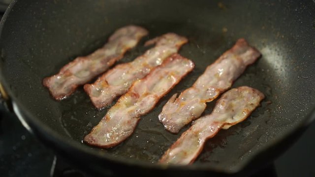 slow-motion of bacon strips being cooked in frying pan