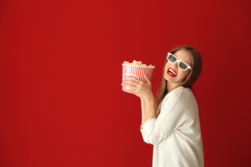 Beautiful young woman with cup of popcorn and 3D cinema glasses on color background