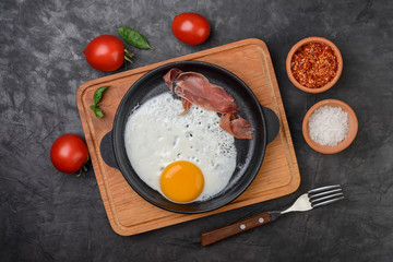 Fried egg in a pan with bacon and  tomatoes  on  black background, top view.