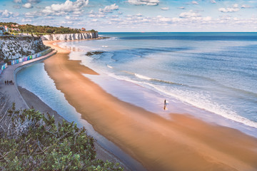 Low tide at Stone Bay, Broadstairs, Kent as summer turns to autumn, a lone surfer walks on the beach and a family on the promenade along side the beach huts and white cliffs. - obrazy, fototapety, plakaty