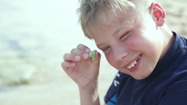 Closeup portrait of happy cheerful smiling kid holding small alive crab in hand. Boy having fun at sunny summer sandy beach during vacations. Real time full hd video footage.