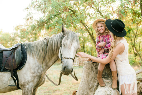 Beautiful young girl with blond hair in a suede jacket with fringe with little sister in a straw hat and checkered vintage dress with a horse in the countryside on a sunny autumn day
