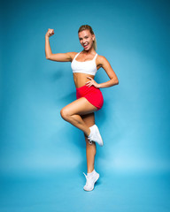 young sports happy sexy blonde girl in red skirt and white top is standing like athlete with strong hands, looking straight and smiling on the blue wall background