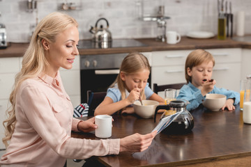 attractive mother having cup of coffee while sitting at kitchen with kids during breakfast