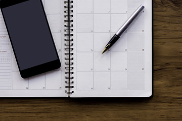 Mockup image of year planner, pen and mobile smartphone with blank screen on wooden table background. Business planning and technology concept. 