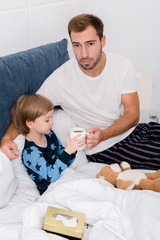 young father giving cup of hot drink to his son in bed and looking at camera