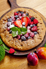 Whole-grain galette with plums and berries on dark background, top view