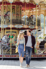 affectionate couple in autumn outfit walking near carousel in amusement park and looking at each other