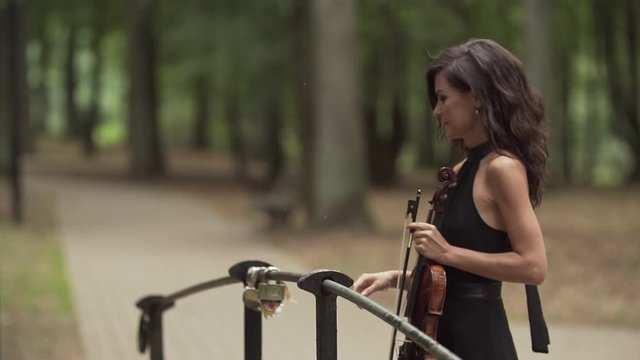 Beautiful girl in black dress with violin in forest bridge. Brunette young violinist in slow motion