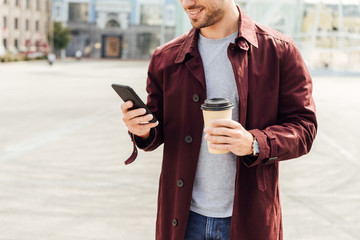 cropped image of man in autumn outfit holding coffee to go and using smartphone in city