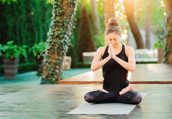 Concentrated girl sitting in lotus pose with hands in namaste and meditating or praying. Young woman with oriental appearance practicing yoga alone on wooden deck in tropical island