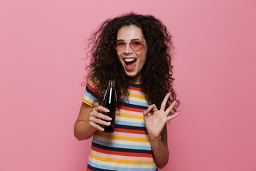 Photo of cheerful woman 20s with curly hair drinking soda from glass bottle, isolated over pink...