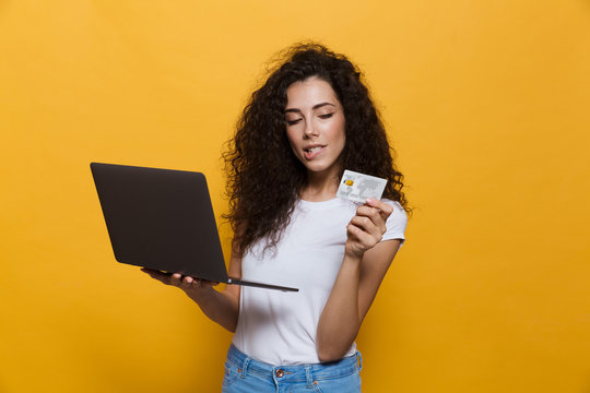 Photo of brunette woman 20s wearing casual clothes holding black laptop and credit card, isolated over yellow background