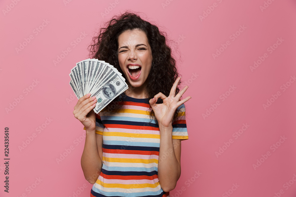 Sticker image of joyous woman 20s with curly hair holding fan of dollar money, isolated over pink background