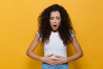 Image of displeased woman 20s with curly hair touching stomach and bending, isolated over yellow...