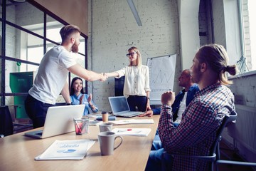 New business partners. Young modern colleagues in smart casual wear shaking hands and smiling while sitting in the creative office.