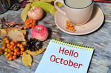 Tag with words hello october on the notebook cup of coffee lying on the books with dry yellow maple leaves and everlasting flowers on background