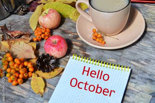 Tag with words hello october on the notebook cup of coffee lying on the books with dry yellow maple leaves and everlasting flowers on background