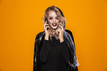 Magician woman wearing black costume and halloween makeup talking on smartphone, isolated over yellow background