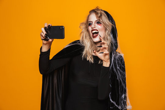Caucasian woman wearing black costume and halloween makeup taking selfie photo on mobile phone, isolated over yellow background