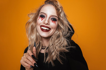 Smiling witch woman wearing black costume and halloween makeup looking at the camera, isolated over...