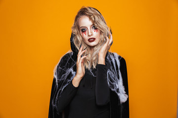 Scary wizard woman wearing black costume and halloween makeup looking at the camera, isolated over yellow background