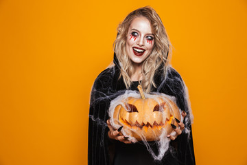 Scary woman wearing black costume and halloween makeup holding carved pumpkin, isolated over yellow...
