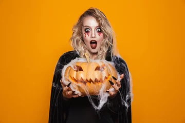 Muurstickers Magician woman wearing black costume and halloween makeup holding carved pumpkin, isolated over yellow background © Drobot Dean