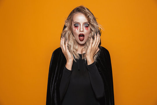 Scary woman wearing black costume and halloween makeup looking at the camera, isolated over yellow background