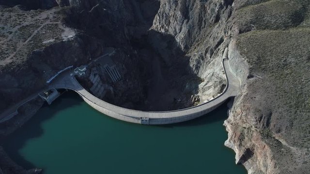 Aerial drone scene of Agua de Toro Dam. Flying over semi circular concrete dam structure camera panning to senital view. Mountains and river canyon at background. San Rafael, Mendoza, Argentina