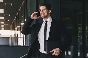 Portrait of employee man dressed in formal suit walking outside glass building, and talking on mobile phone