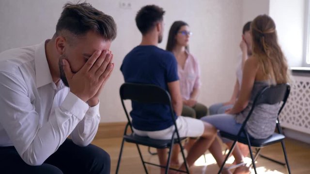 group therapy session, sad man covers his face with hands on background of people sitting on chairs in a circle