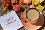 Tag with words hello autumn on the notebook cup of coffee lying on the books with dry yellow maple leaves and everlasting flowers on background