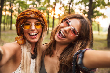 Photo of two cheerful hippie women, smiling and taking selfie while walking in forest