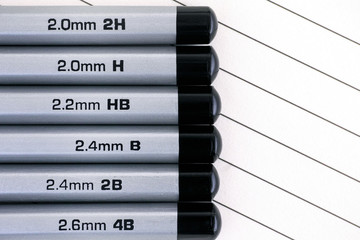 Gray graphite pencils with graphite grading scale in a row on lined paper.